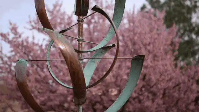 Animated close-up of a Whitaker Studio kinetic wind sculpture spinning in front of blossoming trees