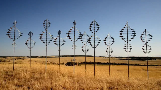 A row of Whitaker Studio kinetic wind sculptures standing in a golden field under a clear blue sky
