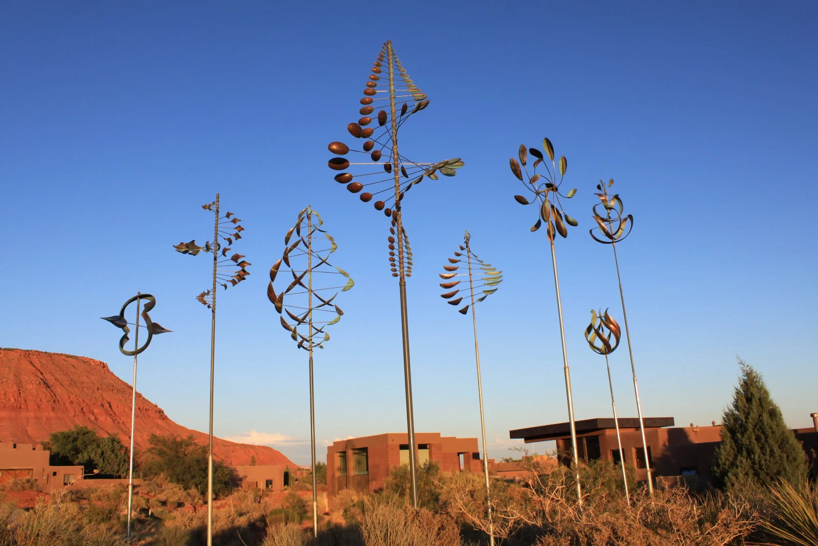 A collection of Whitaker Studio kinetic wind sculptures at sunset against a red rock landscape near modern desert homes
